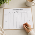 Weekly family planner on a desk with a hand holding a pen, next to a plant and mug.
